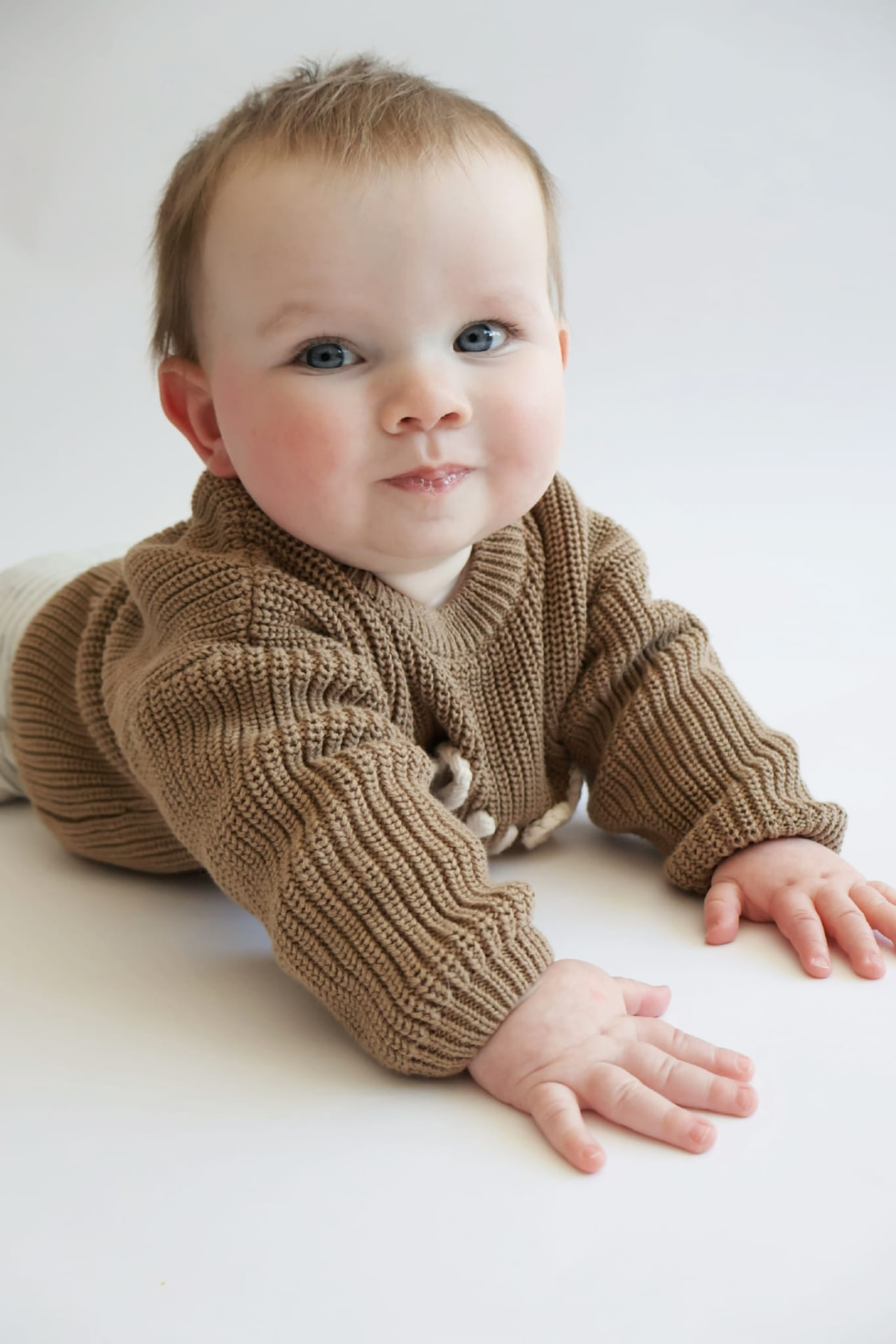 Baby in a brown knit cardigan lying on a soft white backdrop