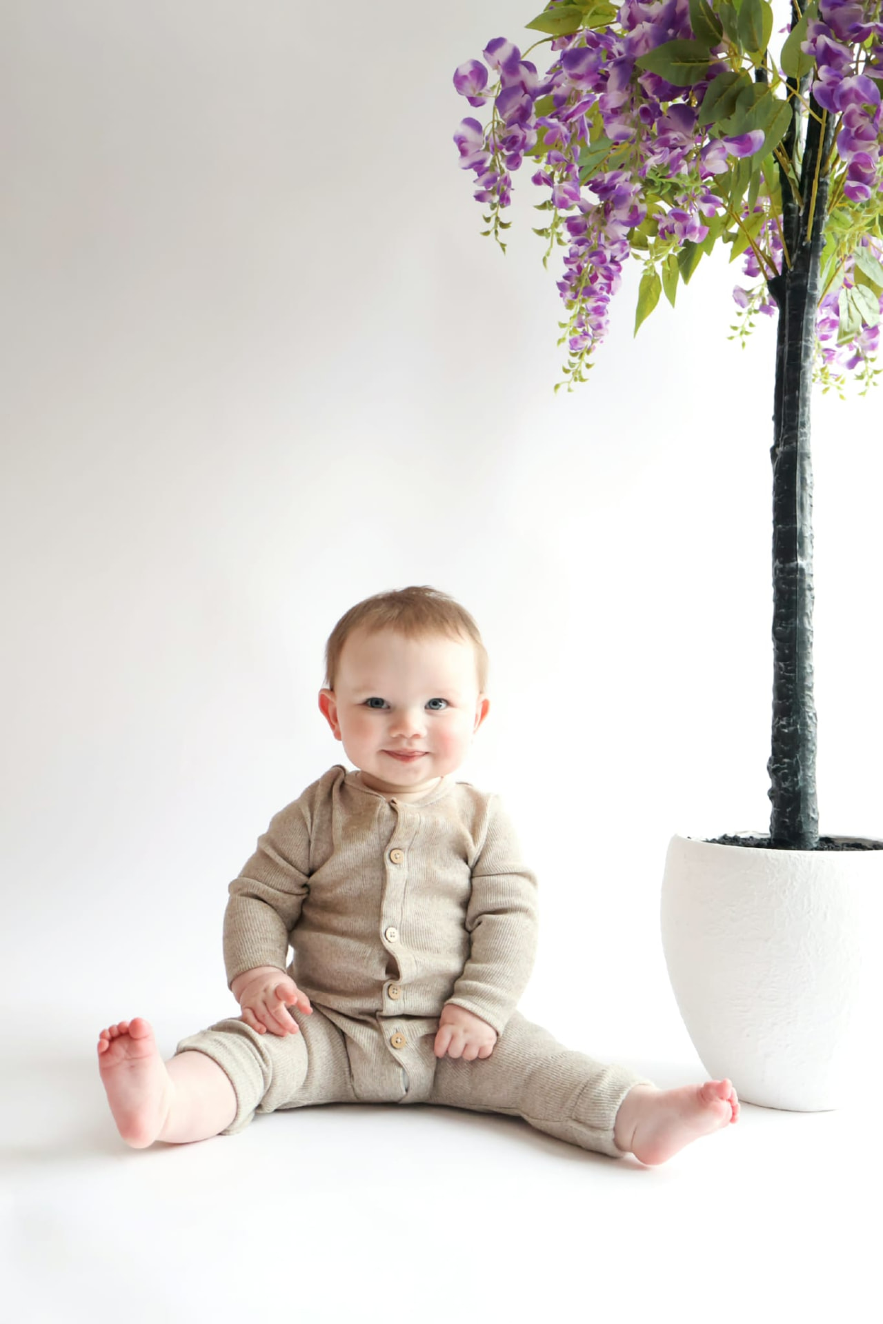 Baby sitting in a beige knit set beside a wisteria tree