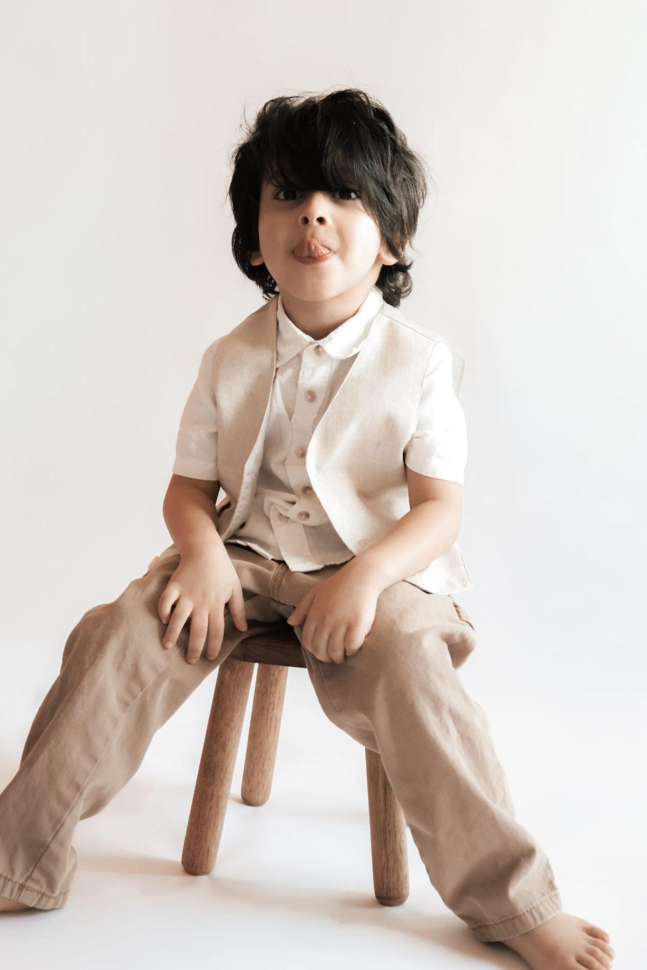 Young boy in a linen vest sitting on a stool making a playful face